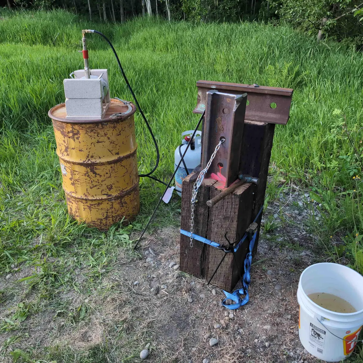 Rail anvil mounted to its stand, with a propane forge set on top of a 55 gallon barrel beside it.