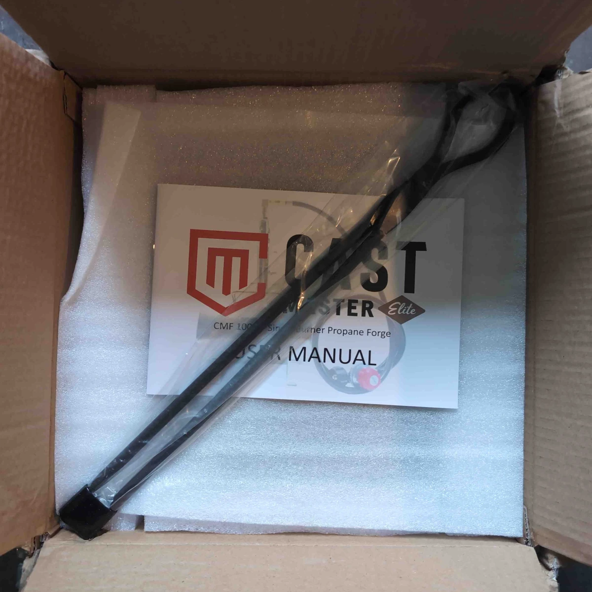 Looking into a box, there is a pair of spindly blacksmith tongs on top of the styrofoam packing.